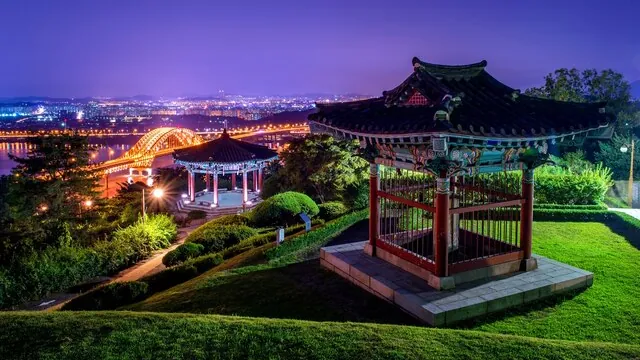 Uma ponte em arco iluminada vista de um parque ribeirinho com pavilhões, à noite.