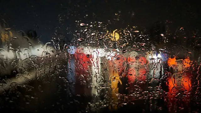 Gotas de chuva numa janela desfocam as luzes de uma rua à noite.