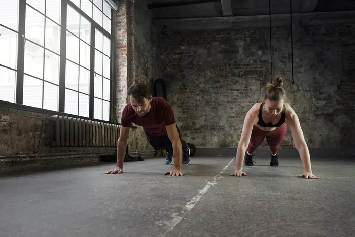 Zwei Athleten machen Liegestütze auf dem Boden einer kahlen Trainingshalle. Burpee Broad Jumps sind eine der Stationen eines HYROX-Rennens.