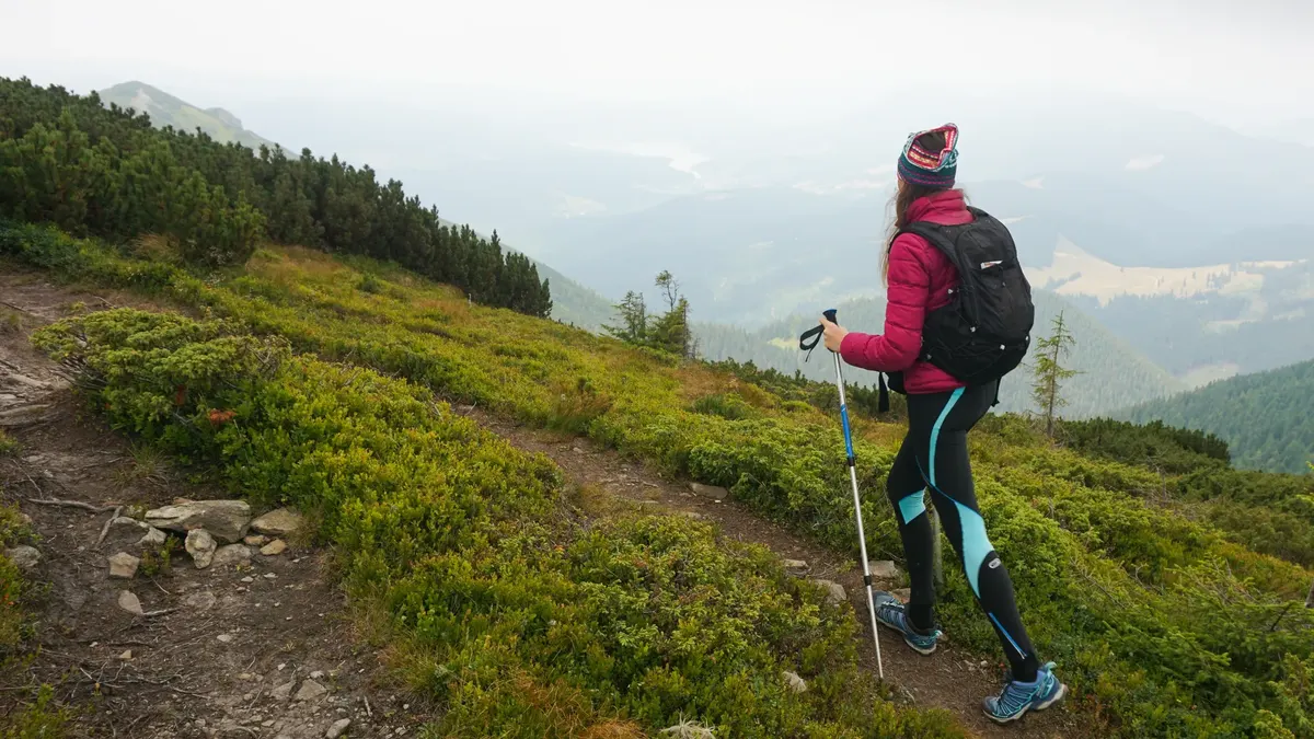 Une randonneuse avec bâtons gravissant une pente herbeuse dans les nuages bas
