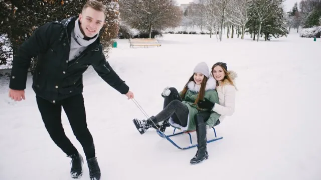 Um homem puxa duas mulheres num trenó por um parque coberto de neve.