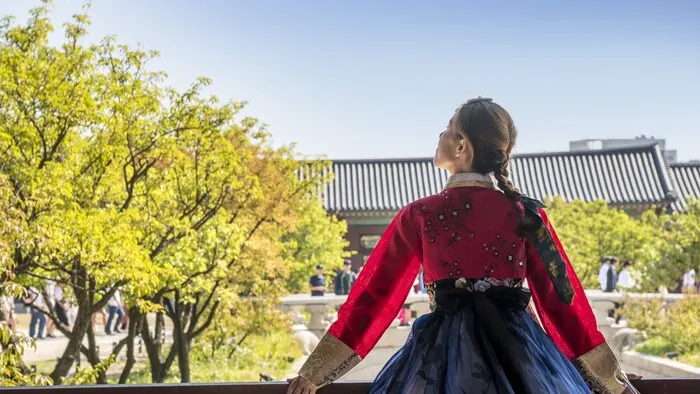 Una mujer con un hanbok rojo y azul mira hacia árboles verdes y un tejado de tejas.