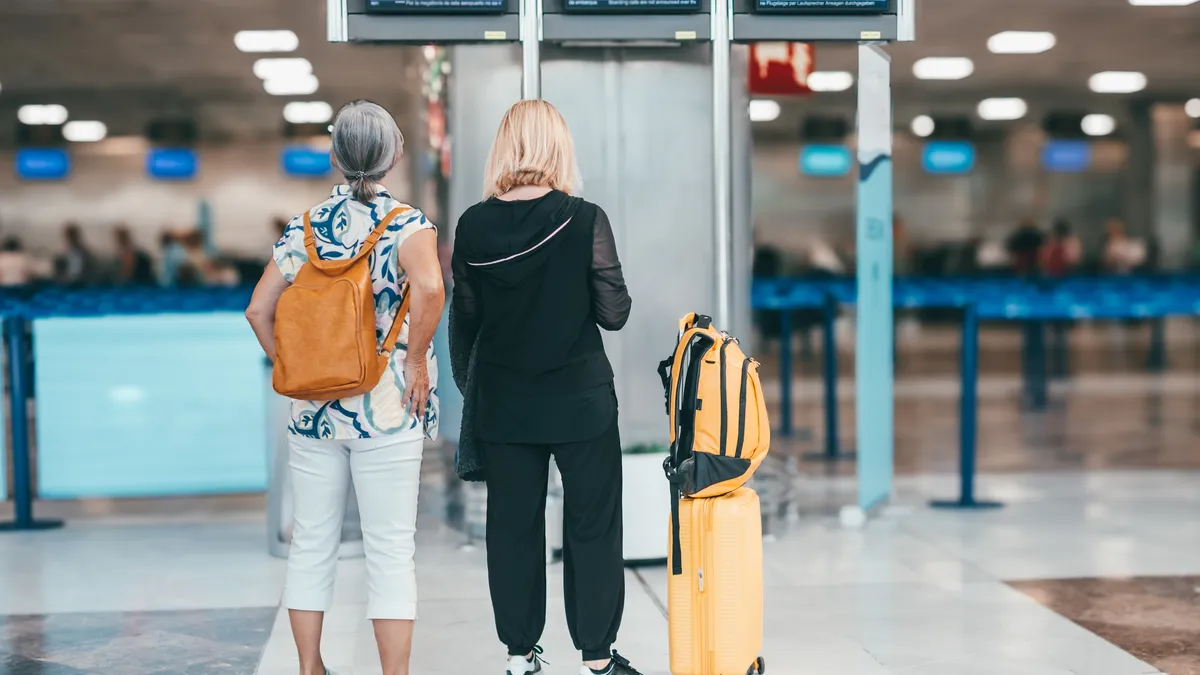 Two travellers with a yellow suitcase and backpack looking towards the departure gates in a bright airport terminal